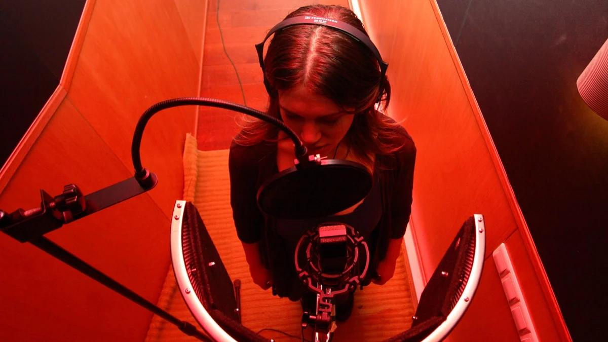 Cantora gravando voz em cabine de estúdio iluminada de vermelho, com microfone condensador e pop filter