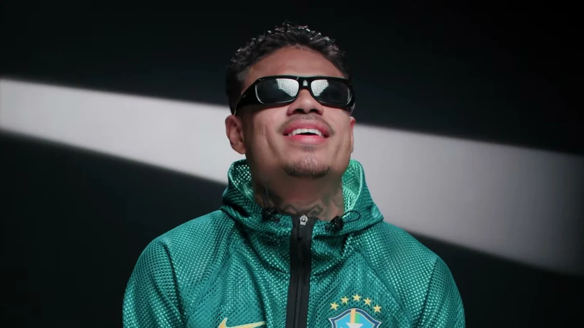 MC Cabelinho de óculos escuros e jaqueta verde da seleção brasileira, sorrindo, em cena do episódio Flavinho de Papai, do Revelando