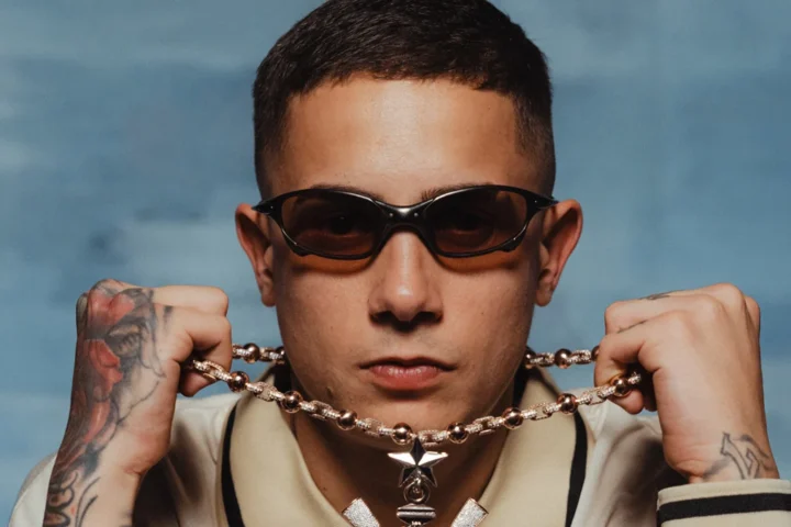 MC Hariel de óculos escuros segurando o cordão diante de um fundo azul