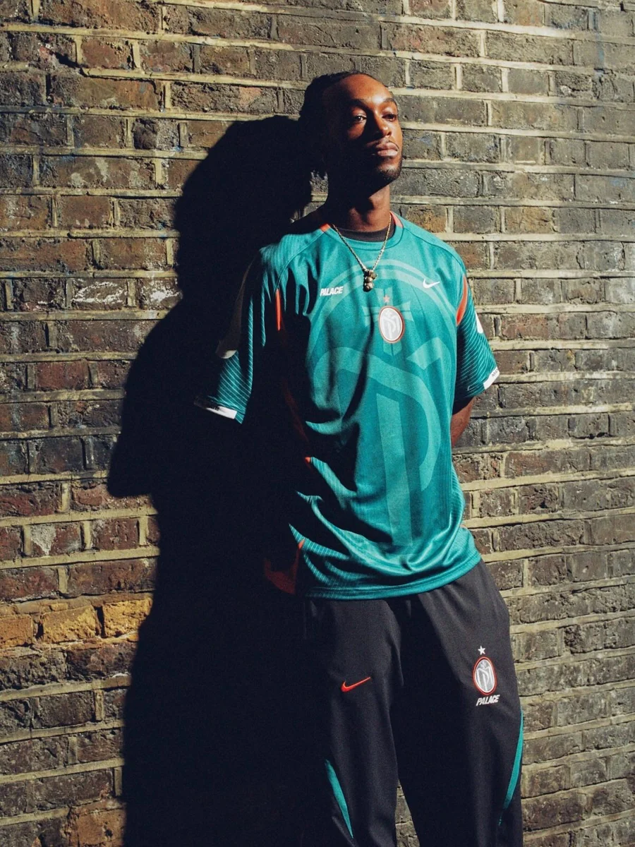 Modelo com camisa azul-petróleo da coleção Palace x Nike Manor Place encostado numa parede de tijolos