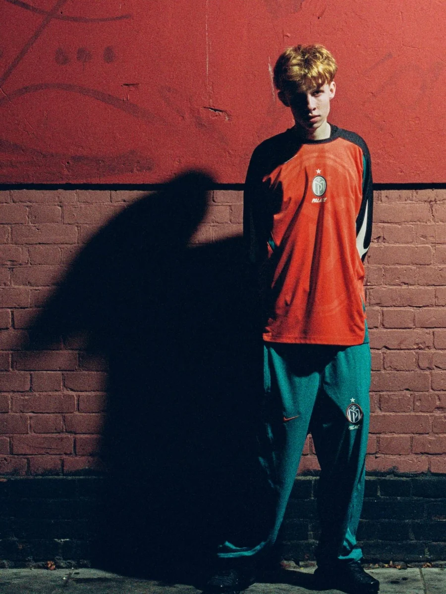 Modelo com camisa laranja e calça azul da coleção Palace x Nike Manor Place